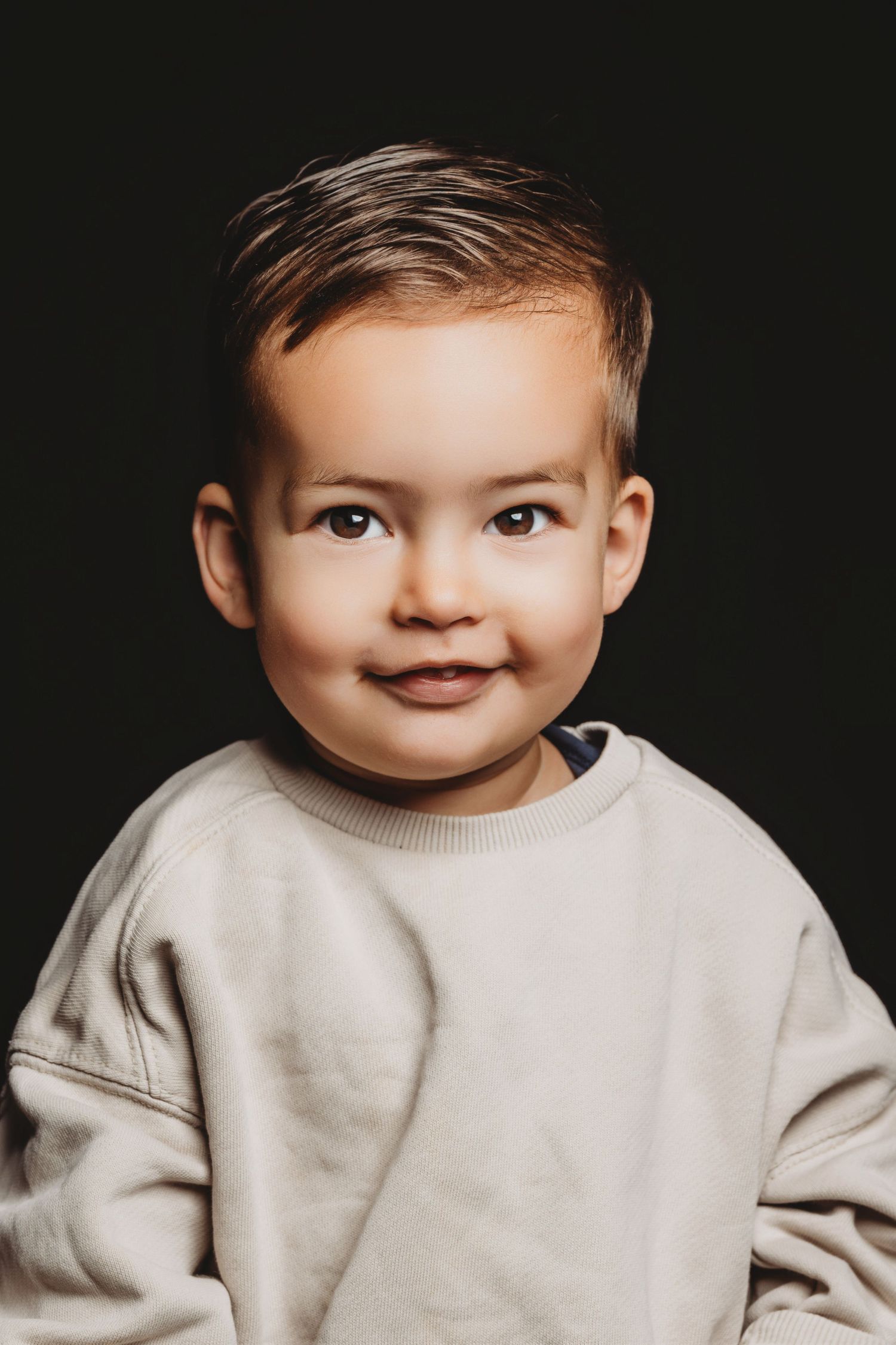 Baby in licht beige trui lacht naar camera tegen donkere achtergrond, kinderfotografie Brabant