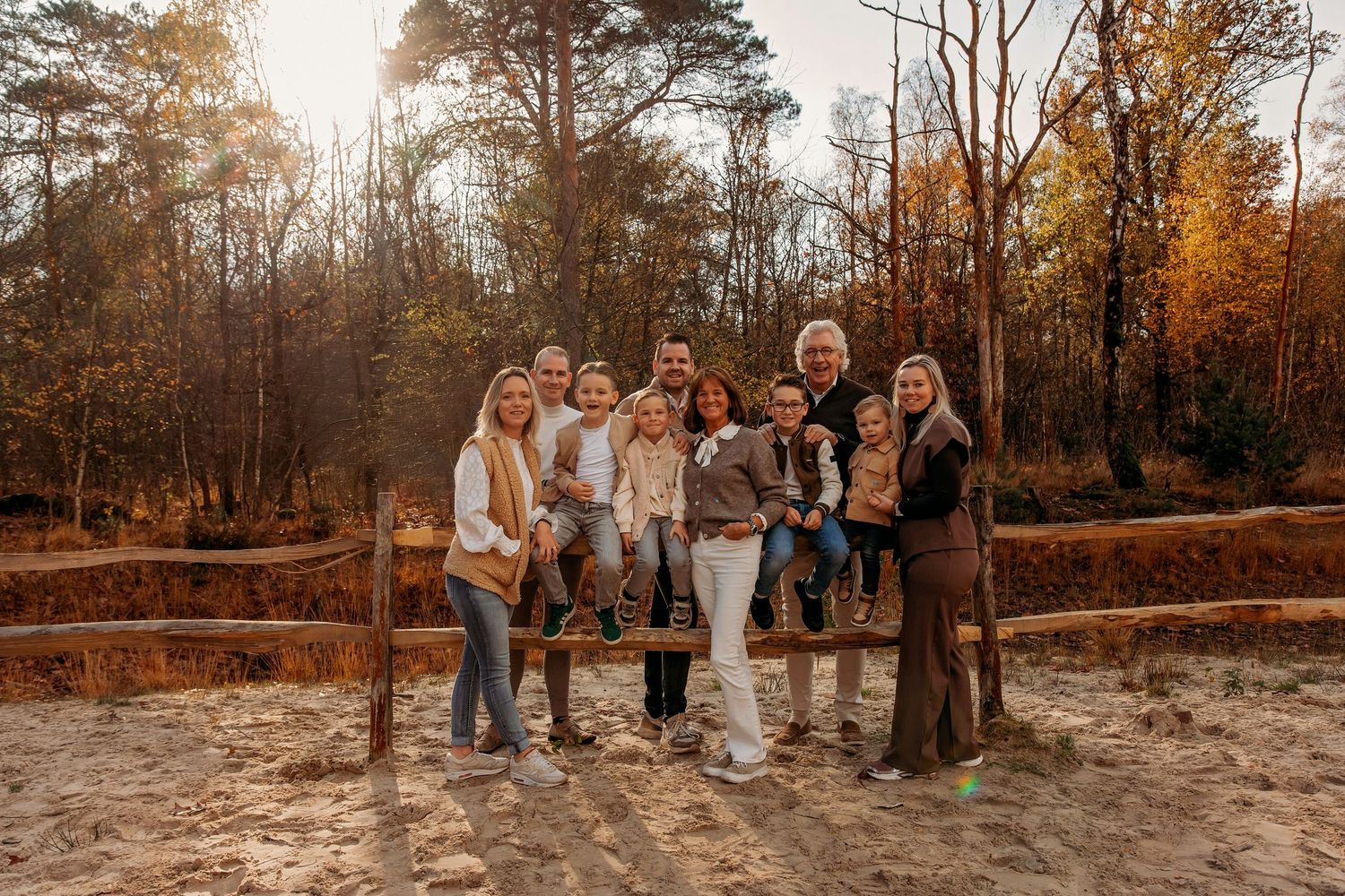 Large family group posing together outdoors near a wooden fence surrounded by autumn trees in golden light.