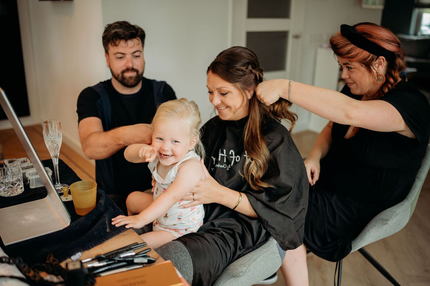 A family with a toddler sits together as a makeup artist styles the mother's hair at a bright indoor setting.