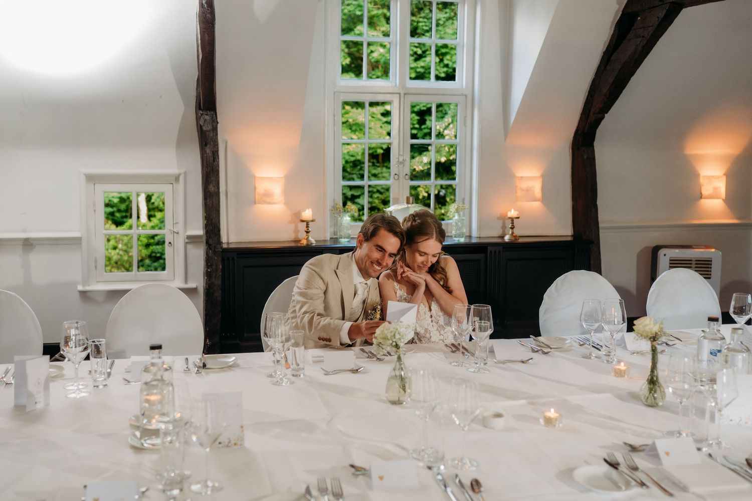 Bride and groom share a tender moment at elegantly set wedding reception table in a bright white venue.