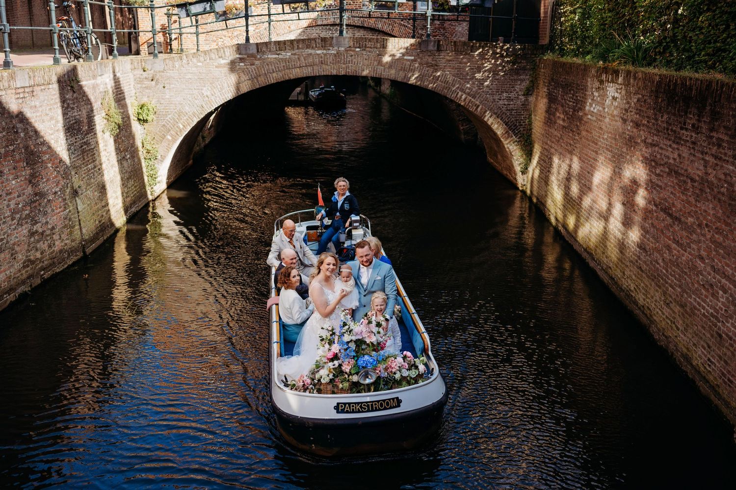 Wedding party on a flower-adorned boat gliding under a brick bridge on a scenic Dutch canal.
