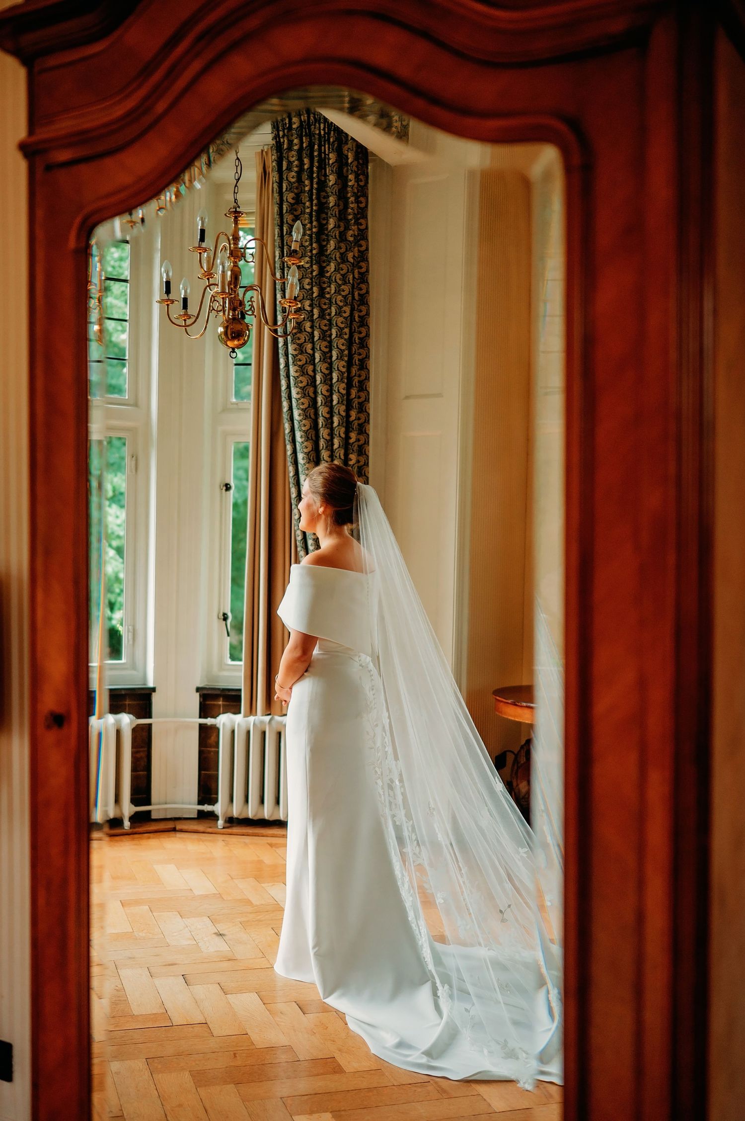 Bride in elegant white off-shoulder gown with long cathedral veil stands by tall windows in ornate room.