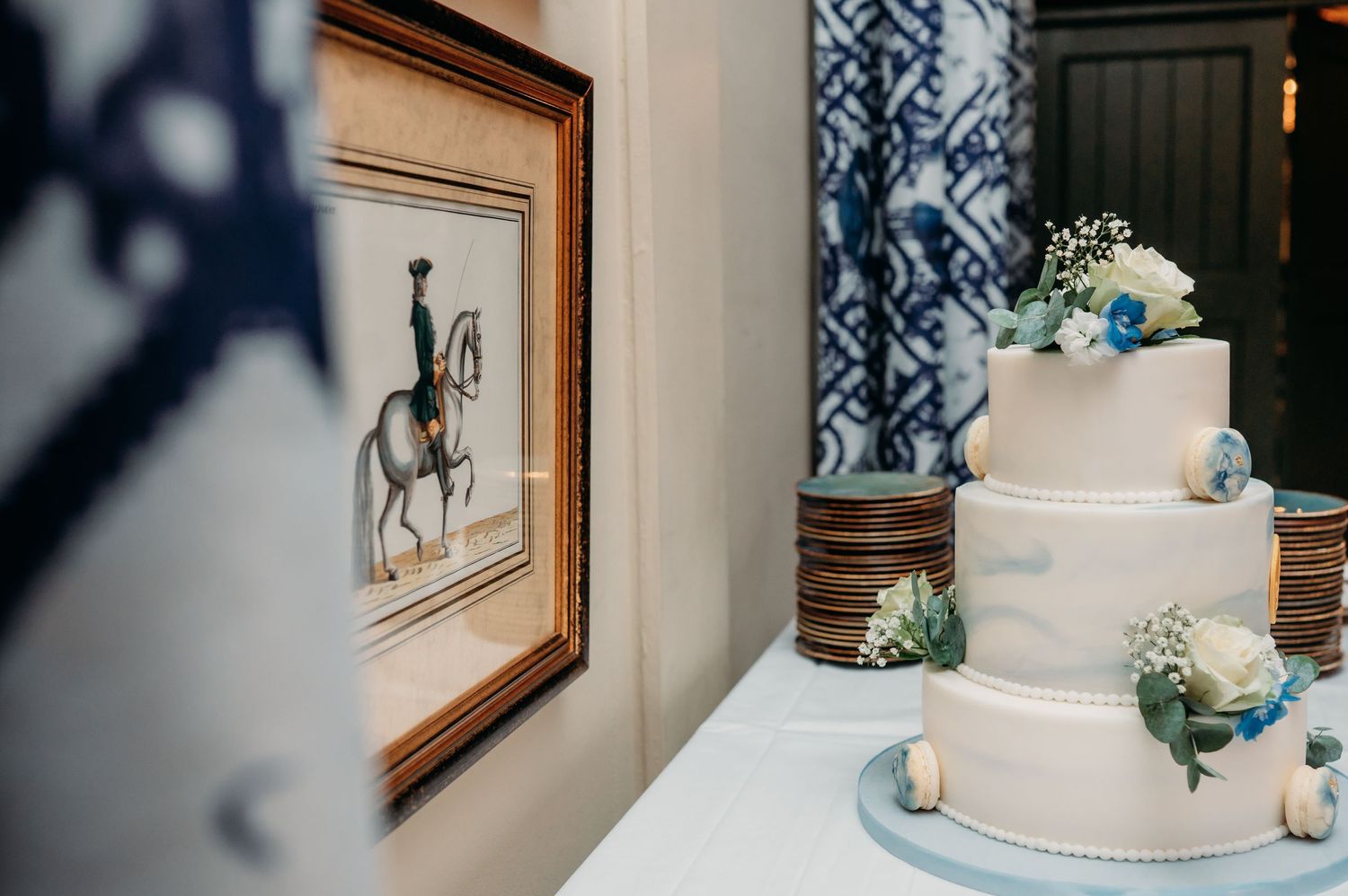 Elegant three-tier white wedding cake with blue floral decorations beside a framed equestrian artwork.