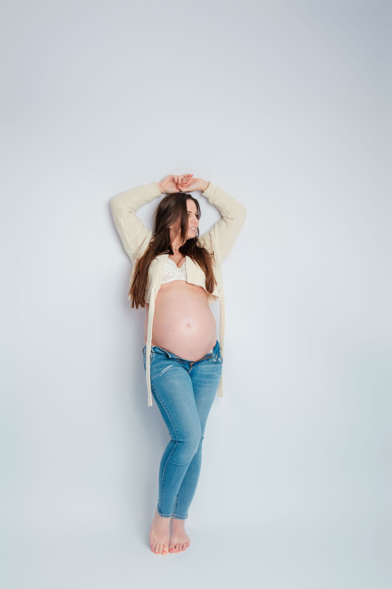 Minimalistische zwangerschapsfotoshoot in jeans en wit top tegen lichte achtergrond, zwangerschapsfotografie Vught, Brabant.