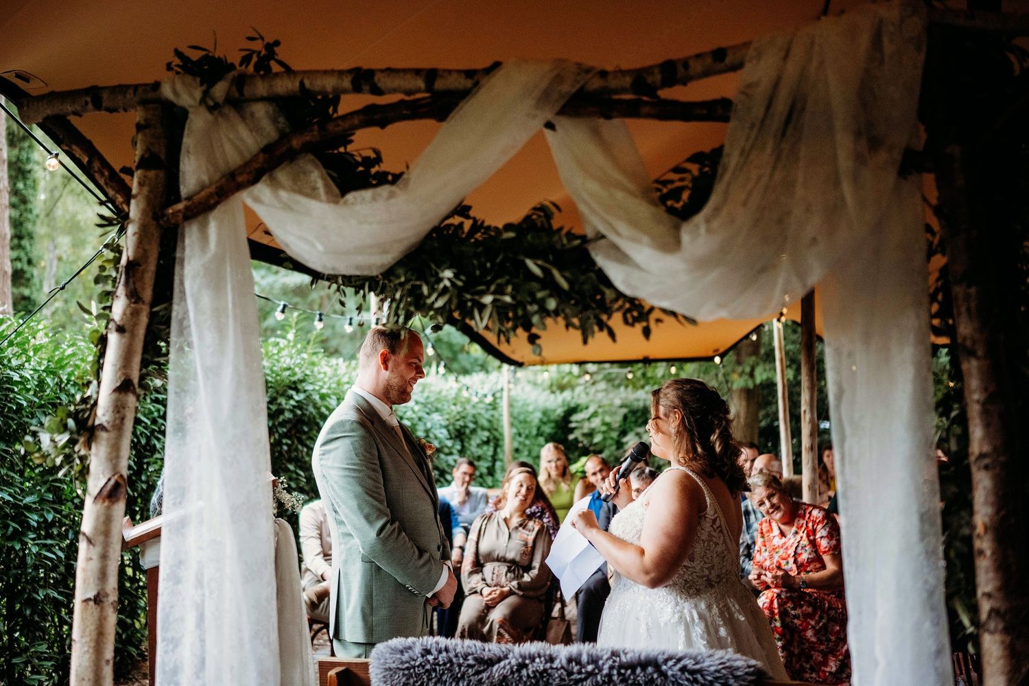 Buiten ceremonie onder houten boog met bloemen, romantische bruidsfotografie in tuinlocatie regio Den Bosch, Vught, Noord Brabant.