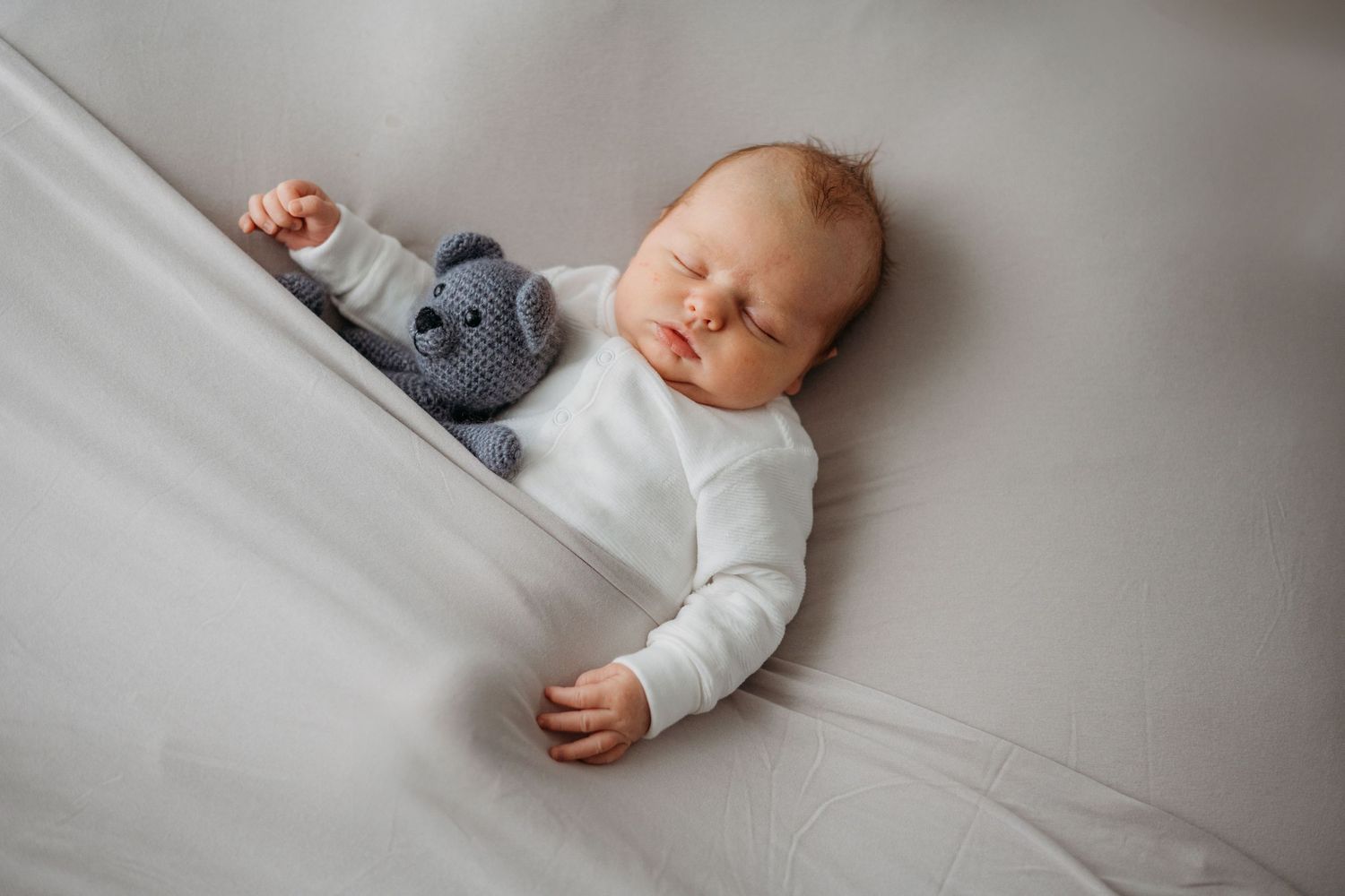 Slapende pasgeborene in witte onesie op lichtgrijze bedding, newbornfotografie Brabant