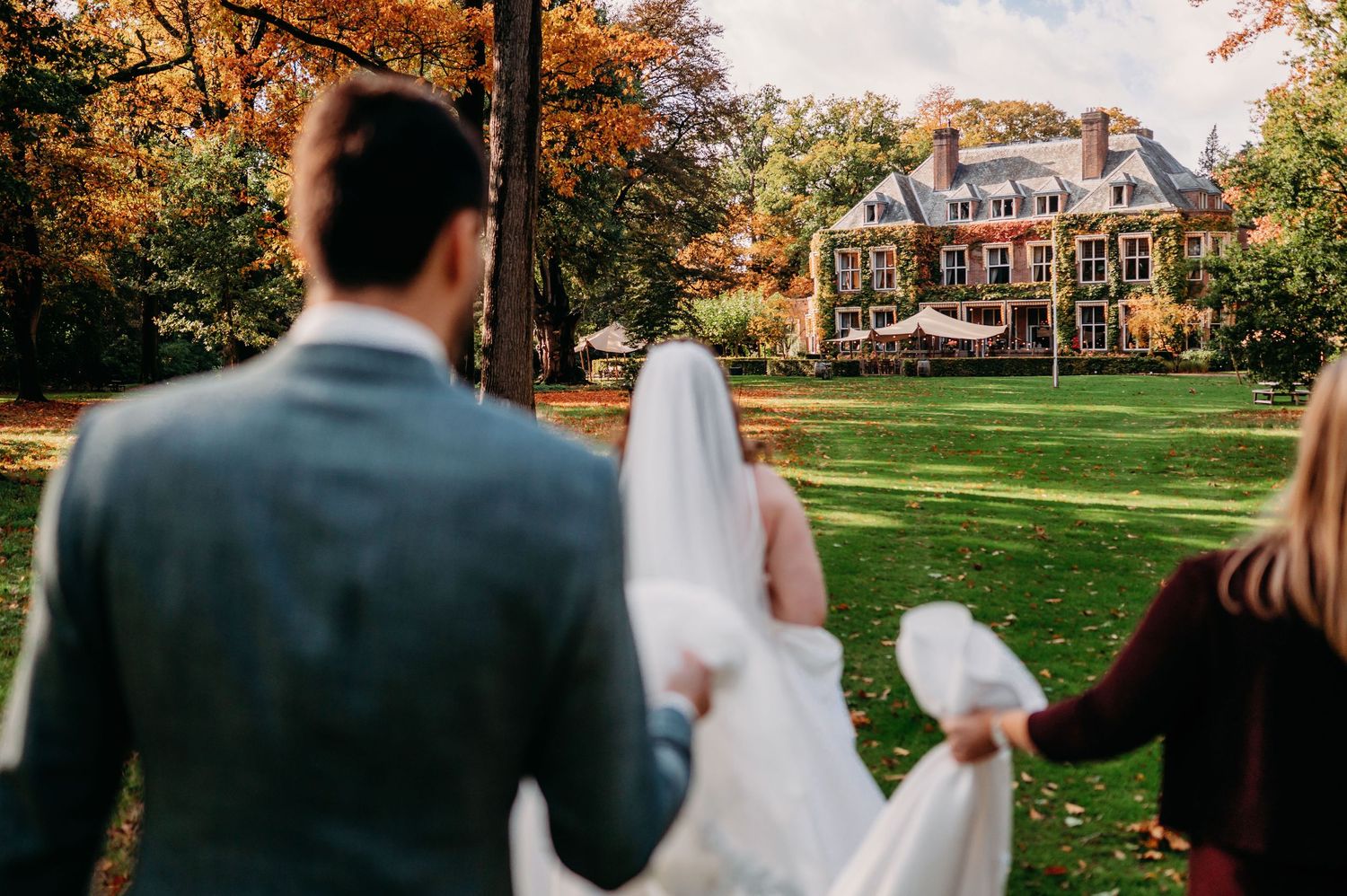A groom and bridesmaid hold the bride's long veil as they walk across a lawn toward a grand manor house in autumn.
