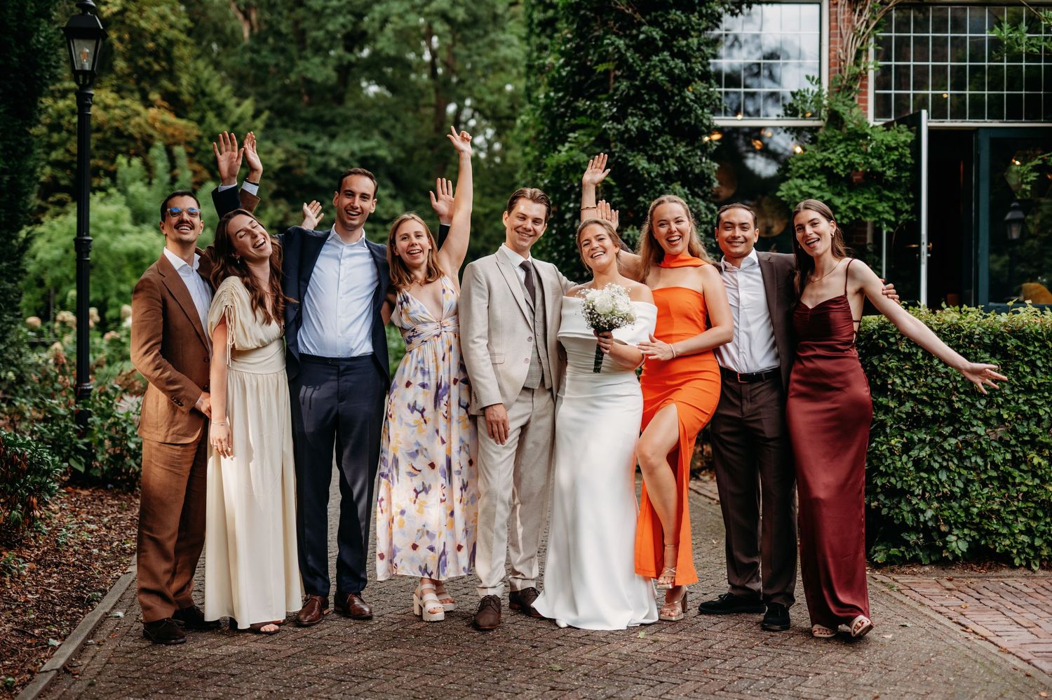 Wedding party of eight celebrating outdoors, bride in white, guests in colorful formal attire, cheering joyfully.