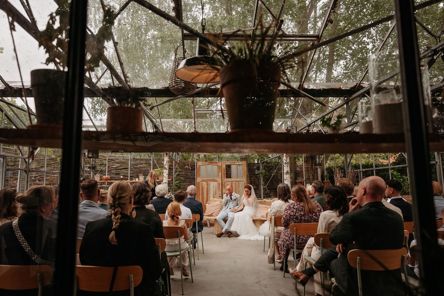 Wedding ceremony inside a stunning greenhouse venue with guests seated among hanging plants and natural light.