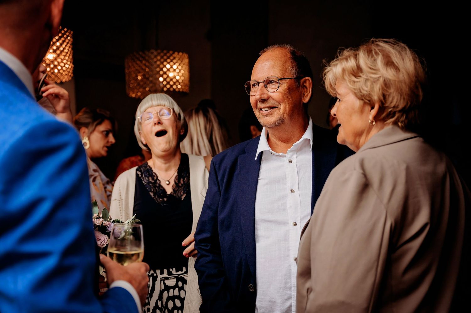 Wedding guests laugh and socialize during a reception, holding glasses of champagne indoors.