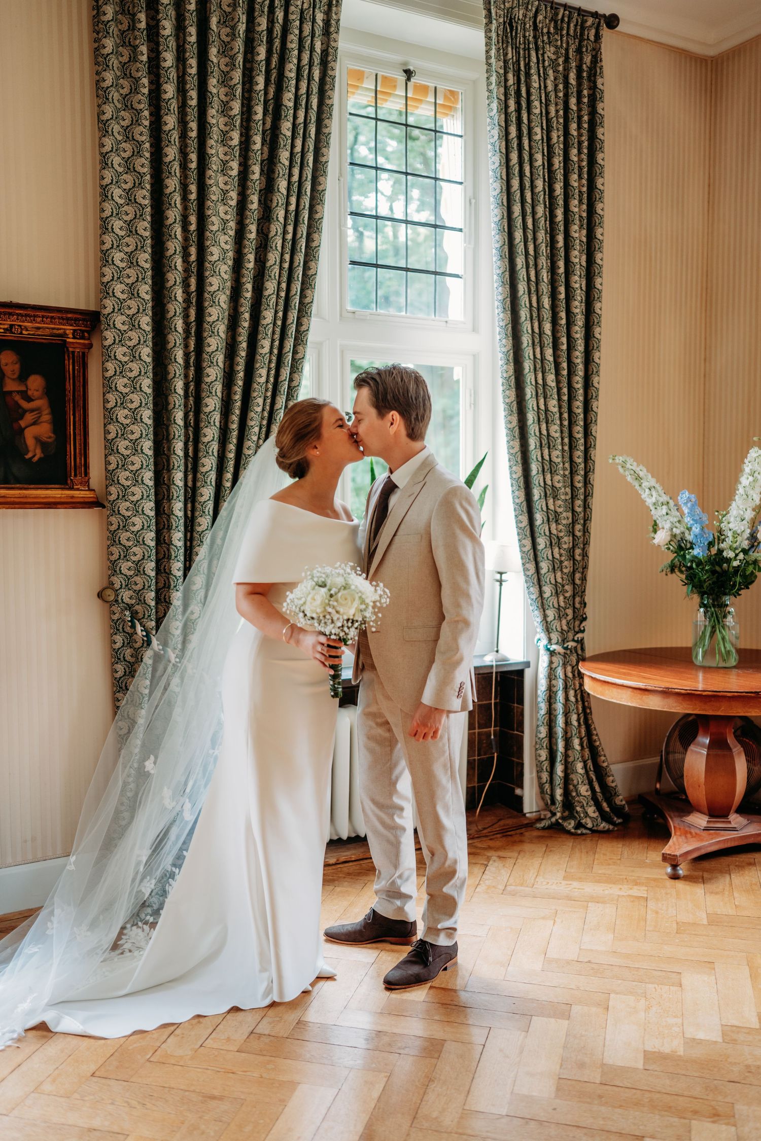 Bride and groom share a kiss in an elegant room with parquet floors, green drapes, and a white floral bouquet.