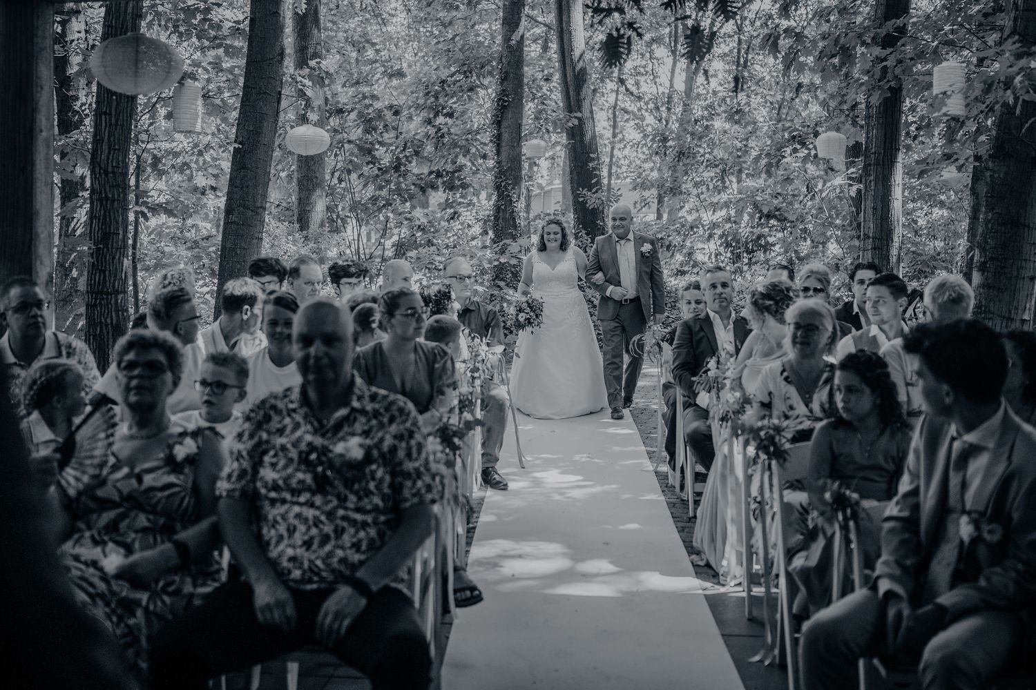 Black and white photo of a bride walking down the aisle at an outdoor woodland wedding ceremony with seated guests.