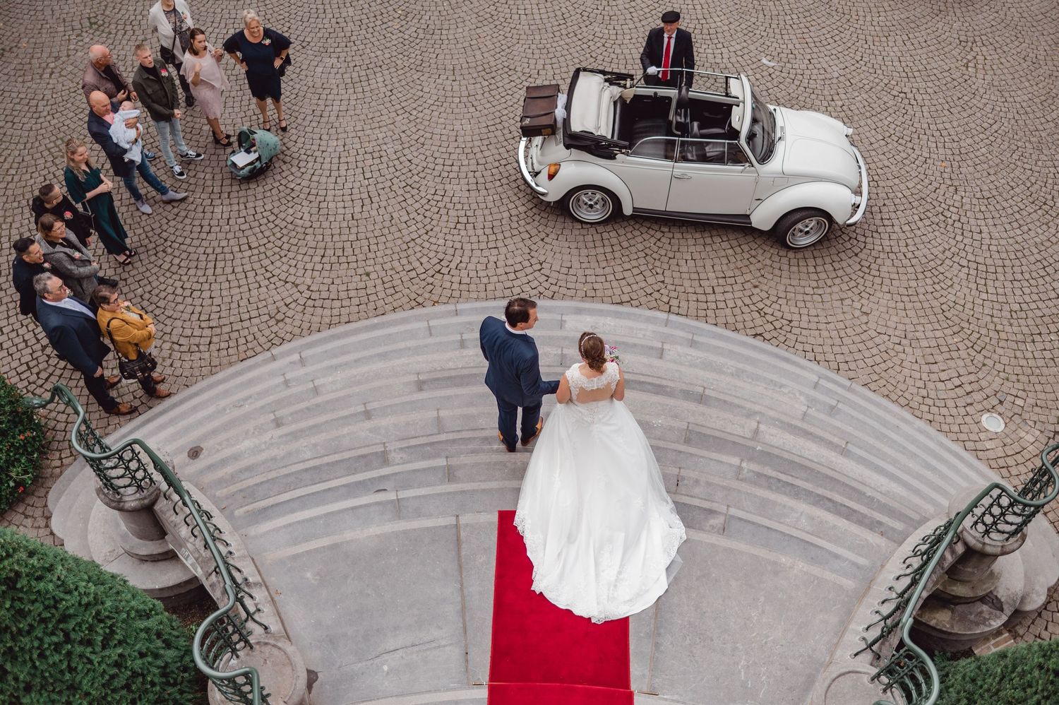 Bruiloftsgasten en vintage witte auto op cirkelvormige stenen oprit voor elegant gebouw, bruidsfotografie Brabant