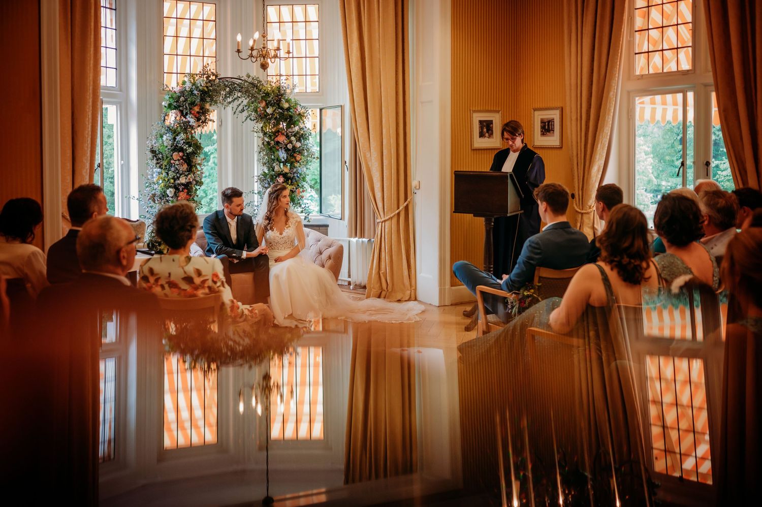 Trouwceremonie in elegante zaal met bloemenboog en gasten, bruidsfotografie Huize Bergen Vught, Brabant.