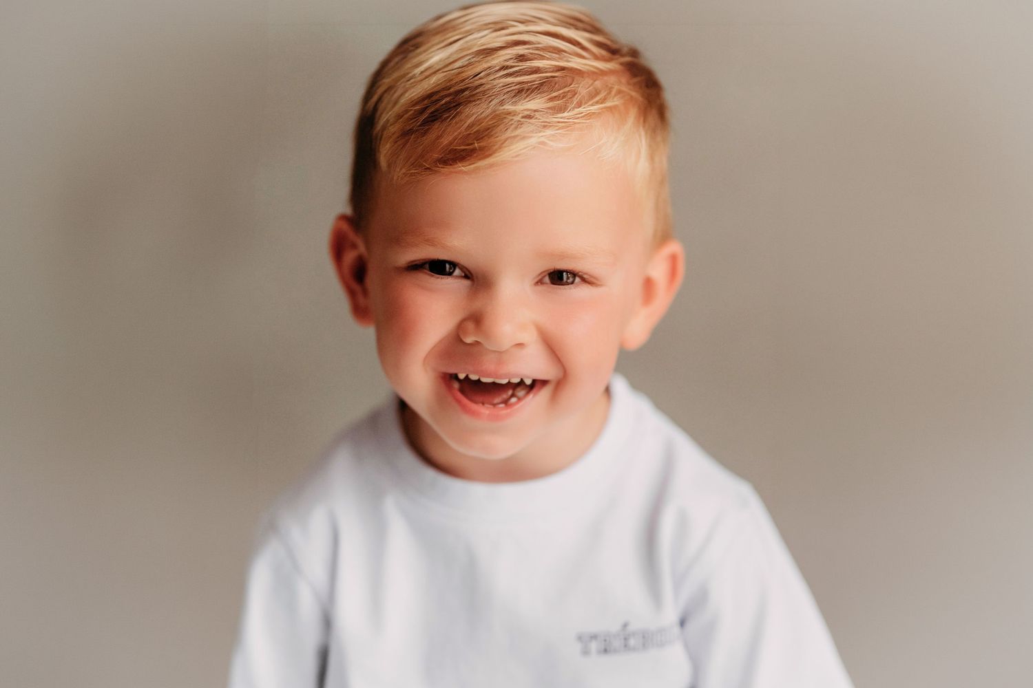 Smiling toddler boy with blonde hair wearing a white sweatshirt against a neutral gray background.