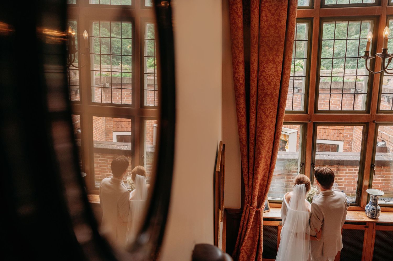 Bride and bridesmaid in white robes looking out large windows with red curtains during wedding preparations.