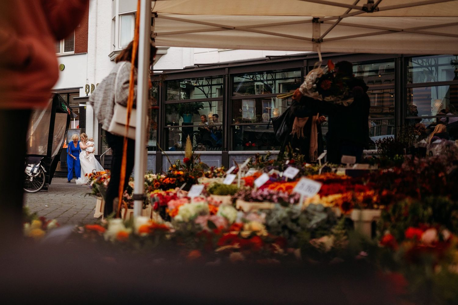Colorful outdoor flower market stall with fresh blooms displayed under a canopy, shoppers browsing vibrant arrangements on a city street.