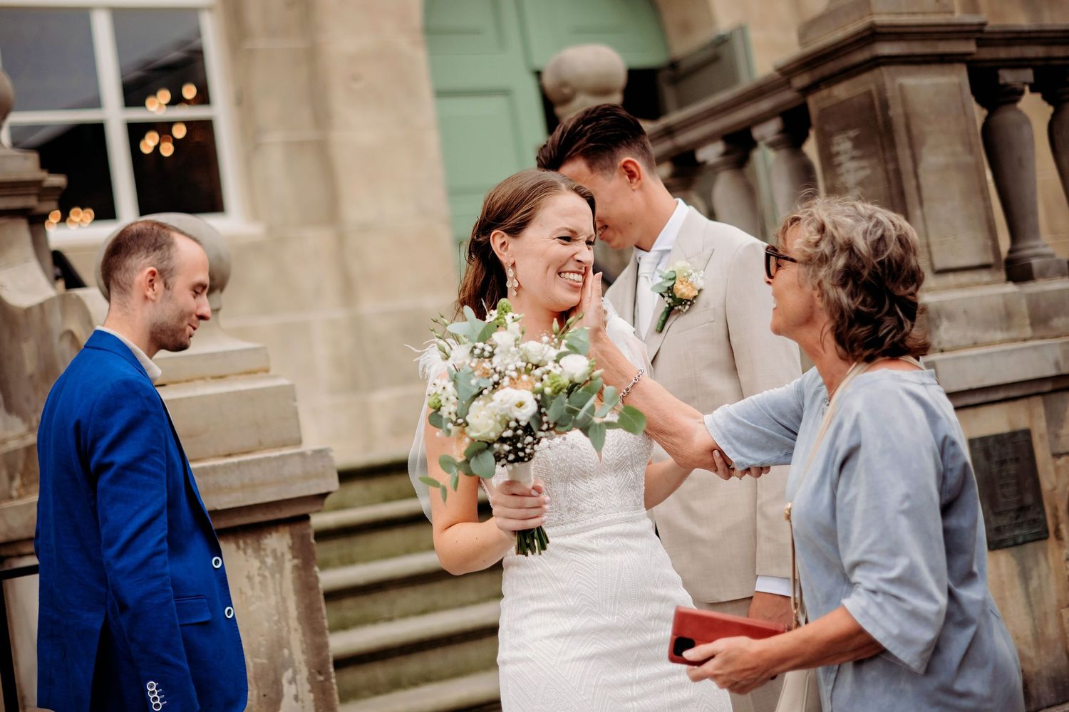 Bride and groom share a joyful moment with guests outside a stone building, bride holding a white floral bouquet.
