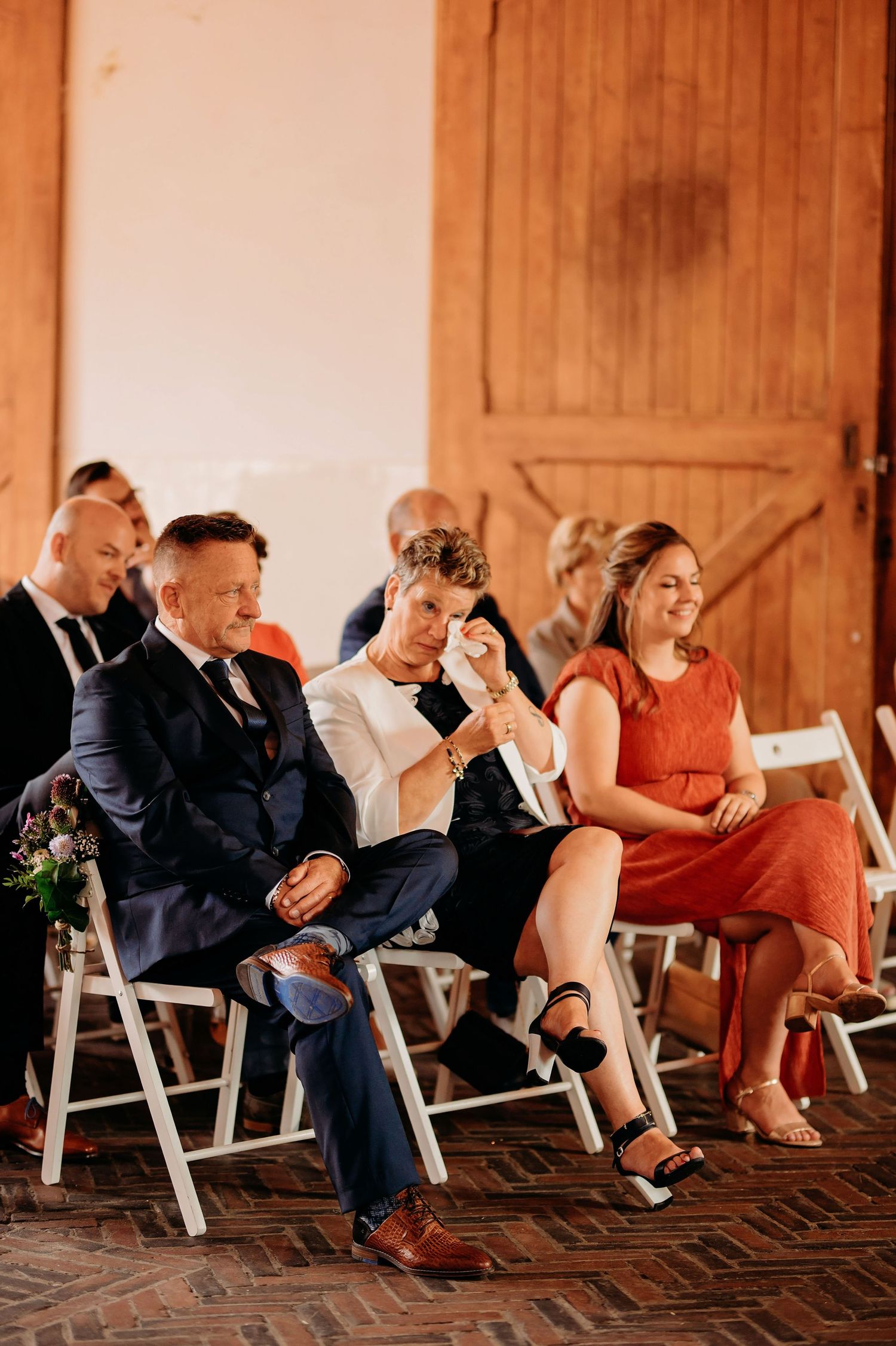 Wedding guests laugh and cry with emotion while seated on white chairs during an outdoor barn ceremony.