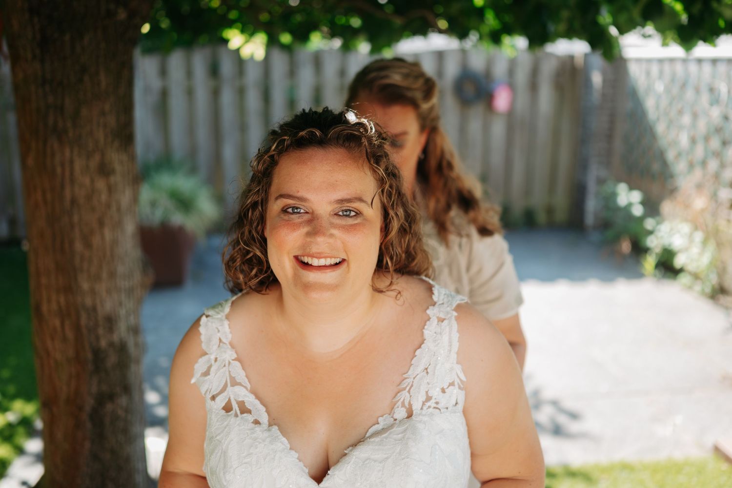 Smiling bride in lace wedding dress outdoors, with bridesmaid helping with dress in sunny backyard.