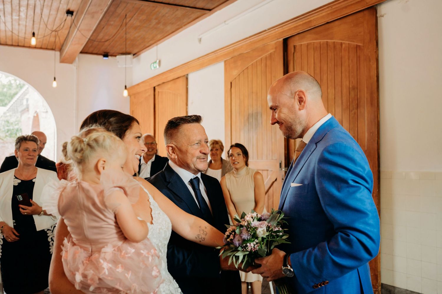 Groom in blue suit greeting bride and young flower girl at wedding ceremony entrance.