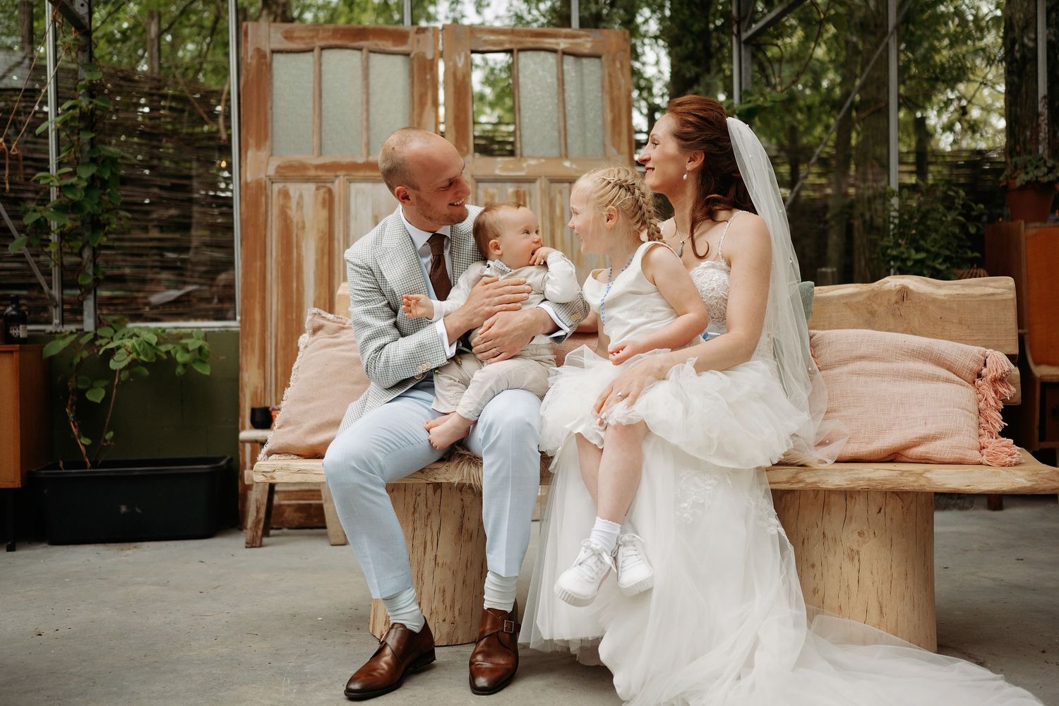 Bride and groom sit on a wooden bench with two young children at their wedding.