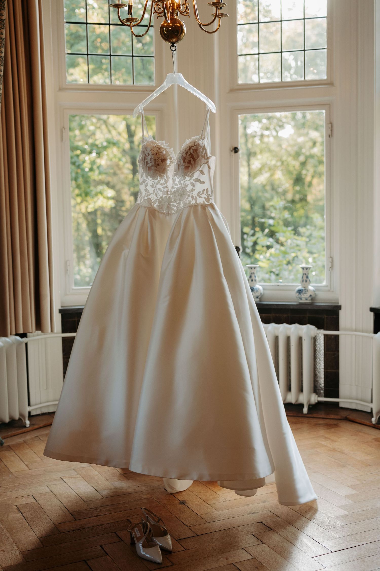 Elegant ivory wedding dress with a lace beaded bodice and satin ballgown skirt hanging by a window, with bridal shoes on the parquet floor.