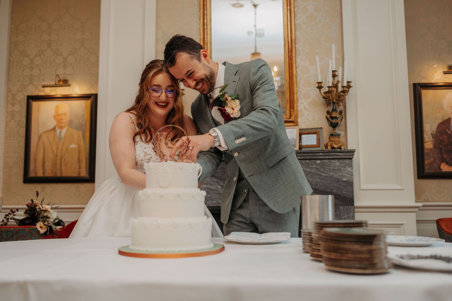 Smiling bride and groom cutting their white tiered wedding cake together in an elegant reception room with framed portraits.