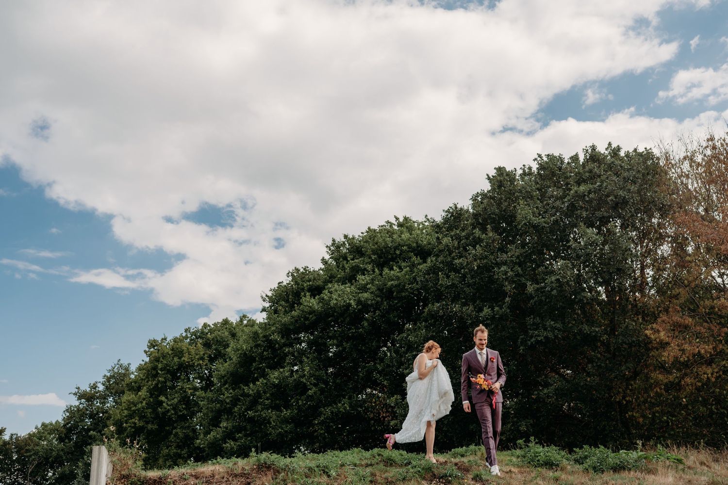 Bruidspaar wandelt samen tijdens hun trouwdag in Brabant onder blauwe lucht, romantische bruidsfotografie