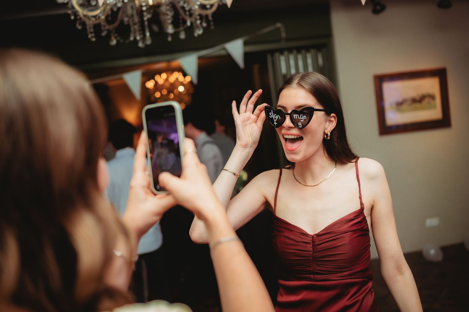 Wedding guest in a red dress poses with oversized sunglasses while a friend photographs her at the reception.
