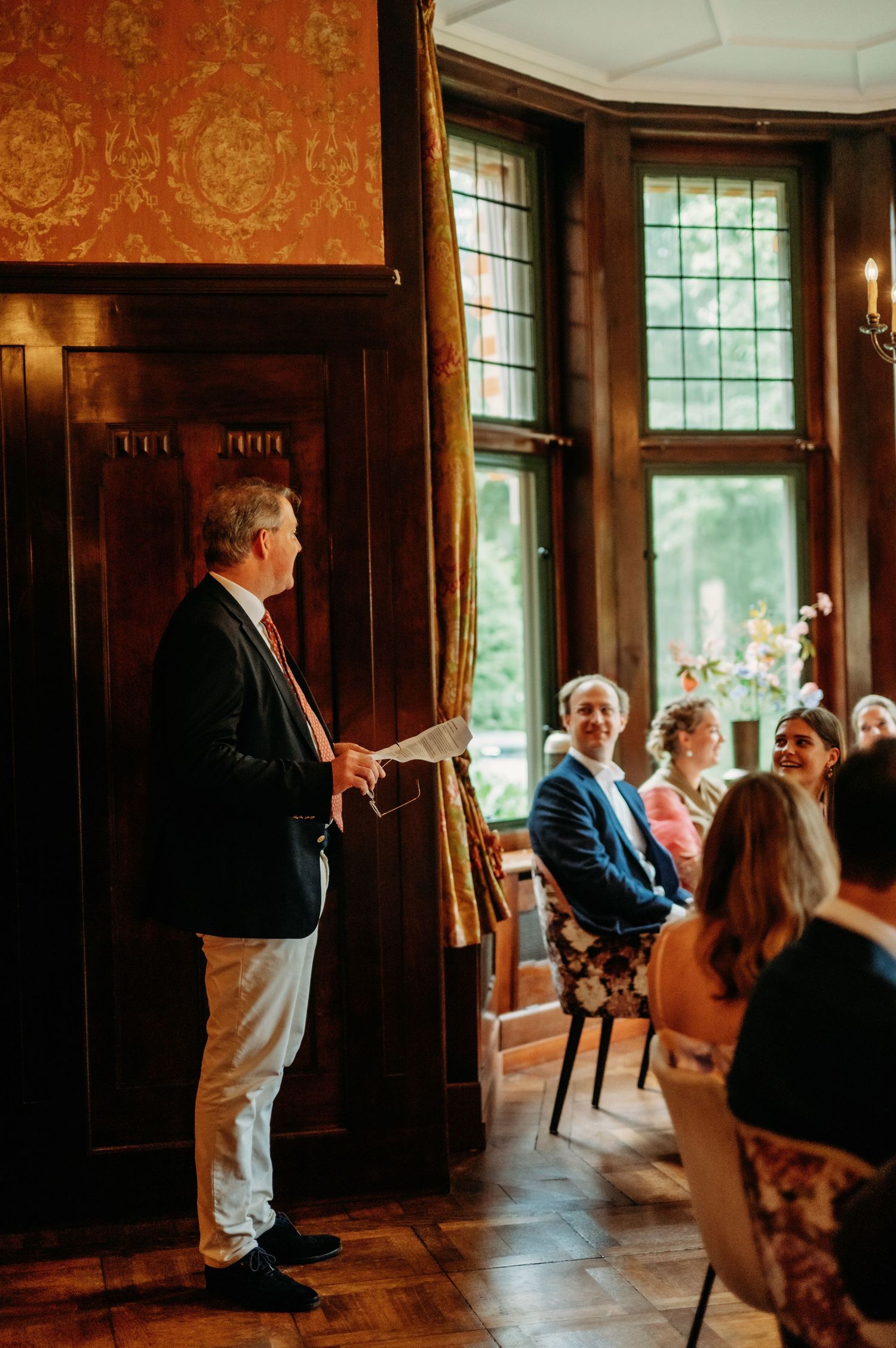 Man in blazer reads speech at elegant wood-paneled wedding ceremony with seated guests smiling in background.