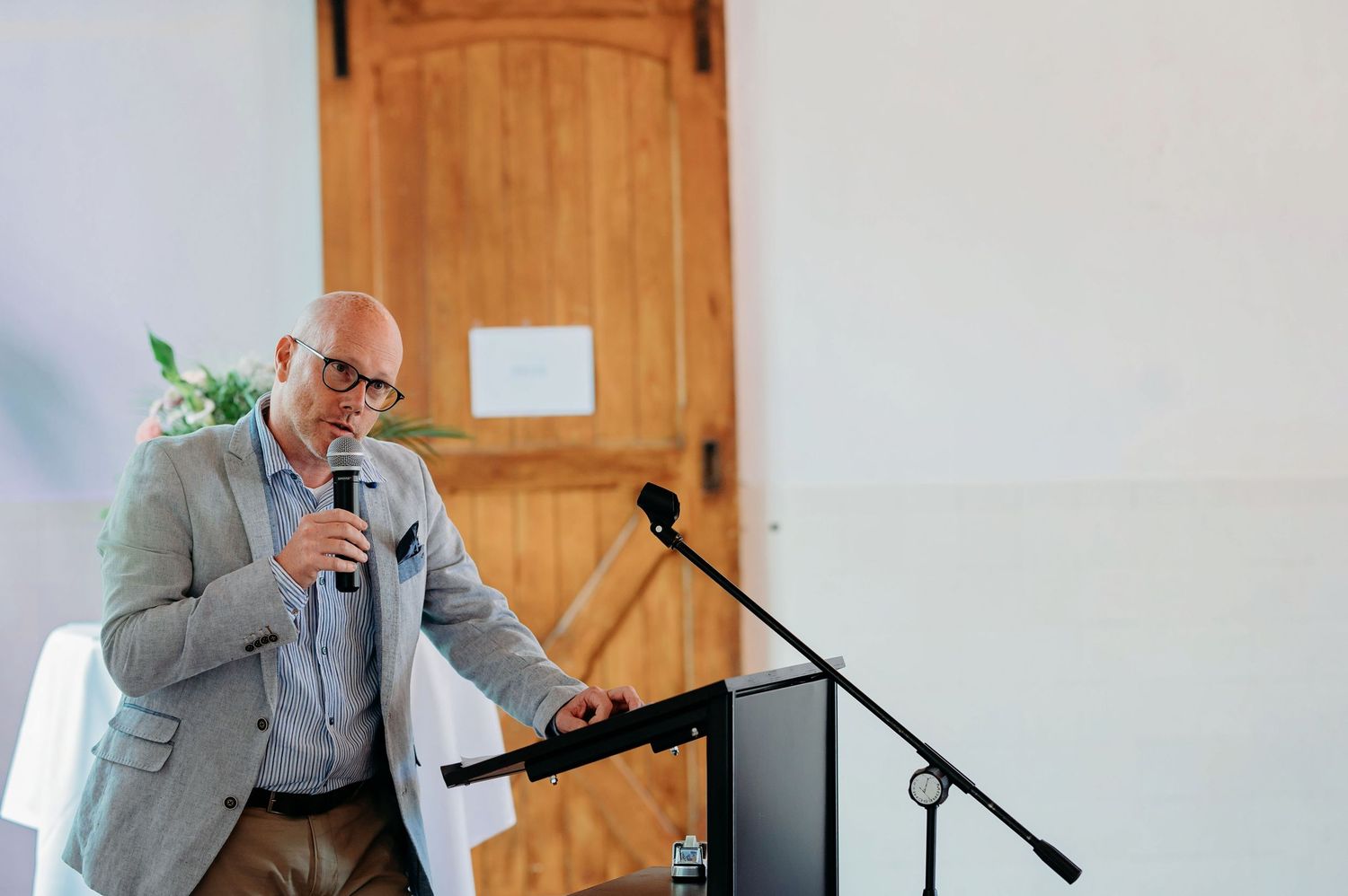 A bald man in a gray blazer speaks into a microphone at a podium during a formal event.