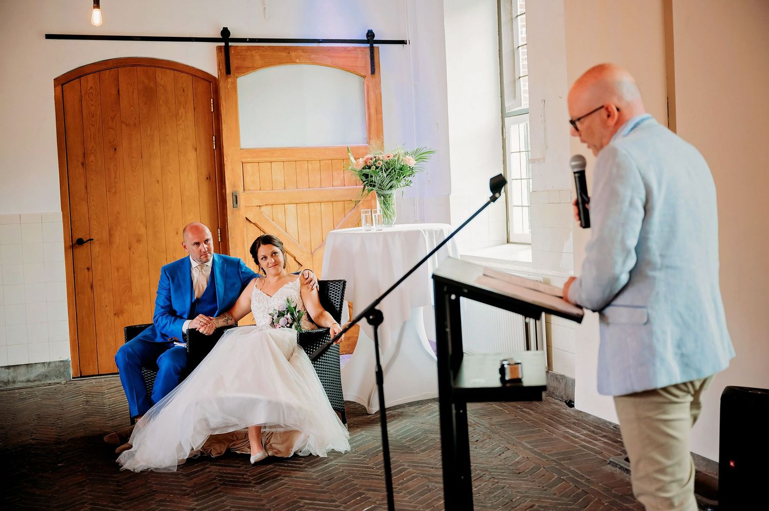 Bride and groom in blue suit laugh during wedding ceremony speech at elegant venue with barn doors.