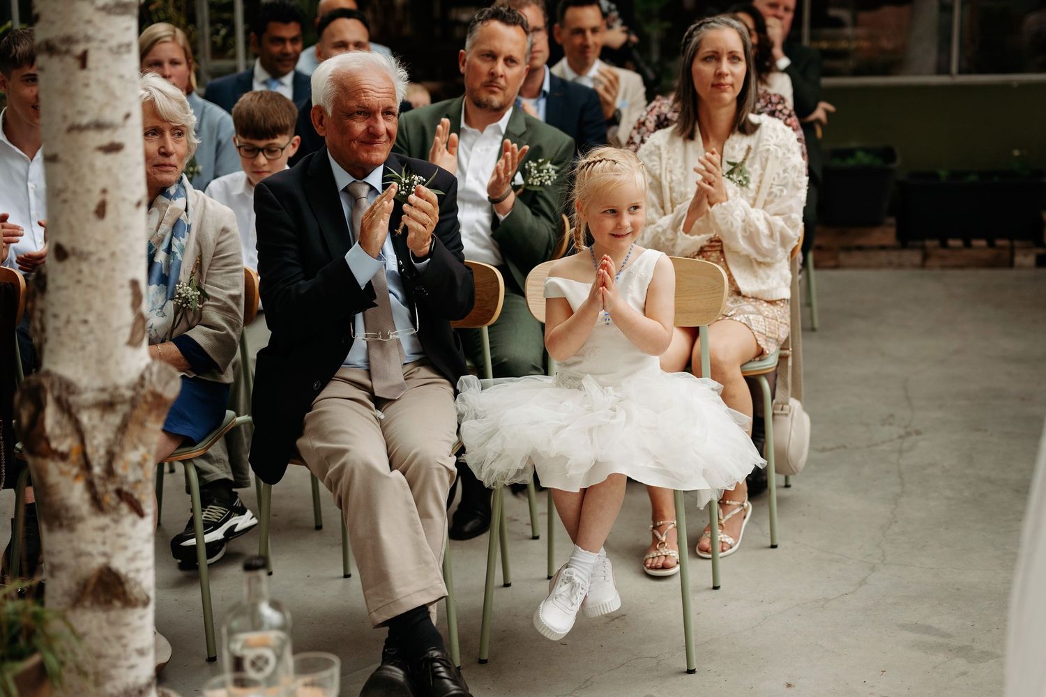 Adorable flower girl clapping with wedding guests during an outdoor garden ceremony celebration.