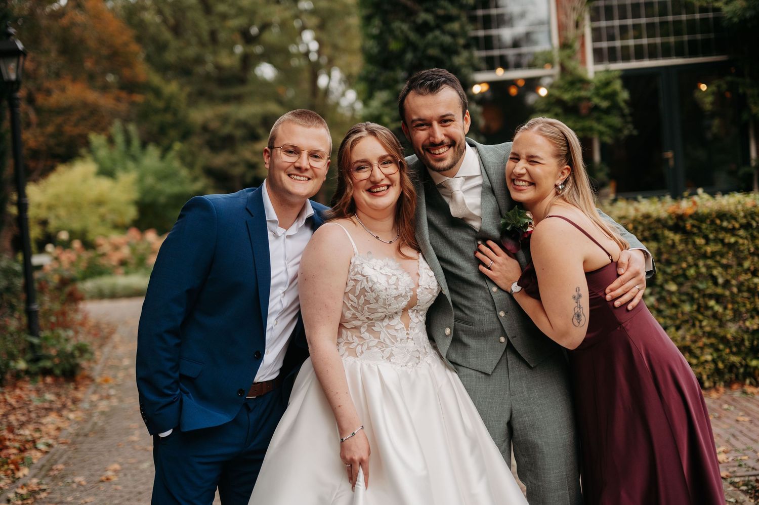 Smiling bride in a white lace wedding gown poses with two groomsmen and a bridesmaid in a burgundy dress in a leafy garden.