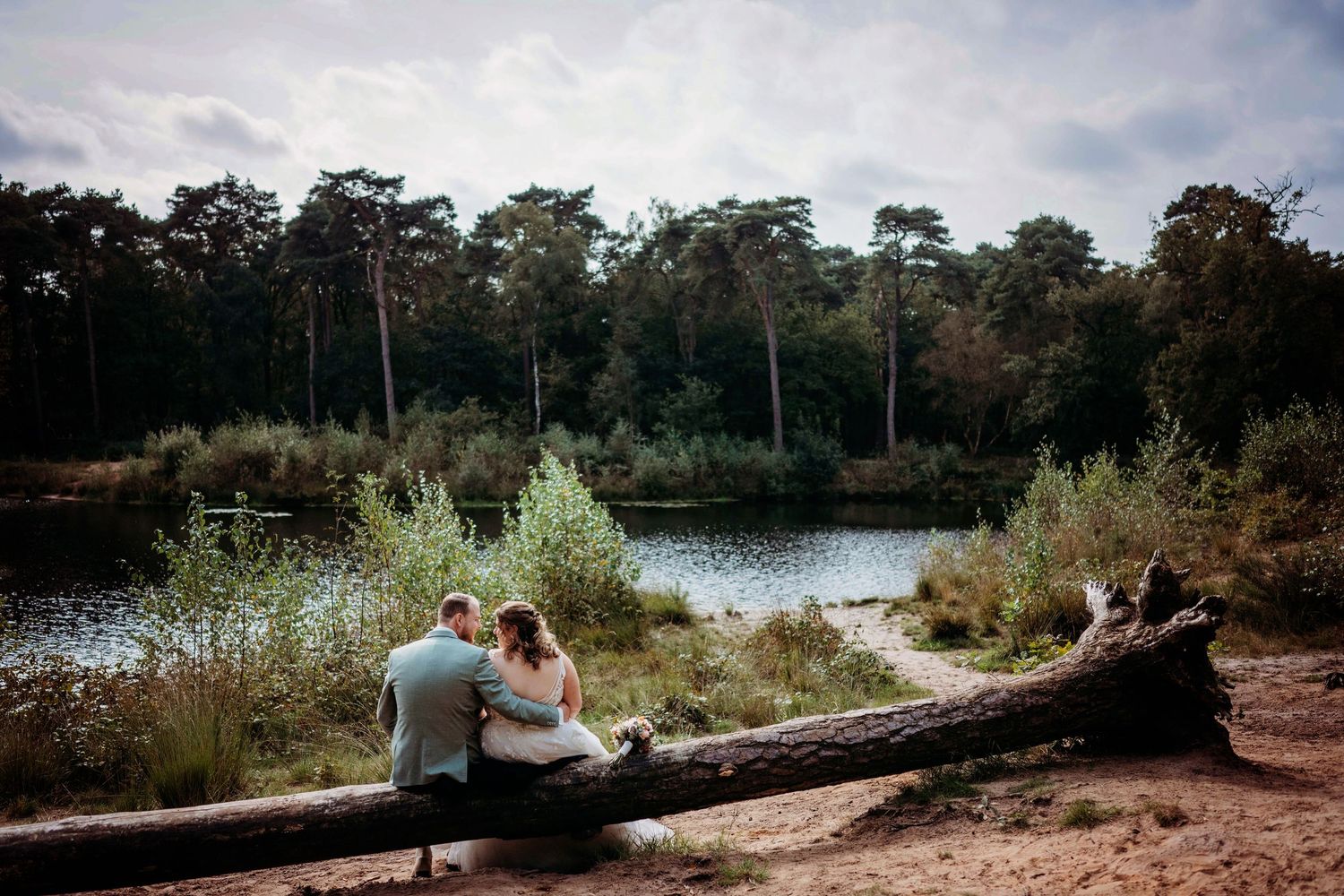 Bruidspaar zit samen aan meer in het bos bij schemer, romantische bruidsfotografie in natuur regio Den Bosch, Vught, Noord Brabant.