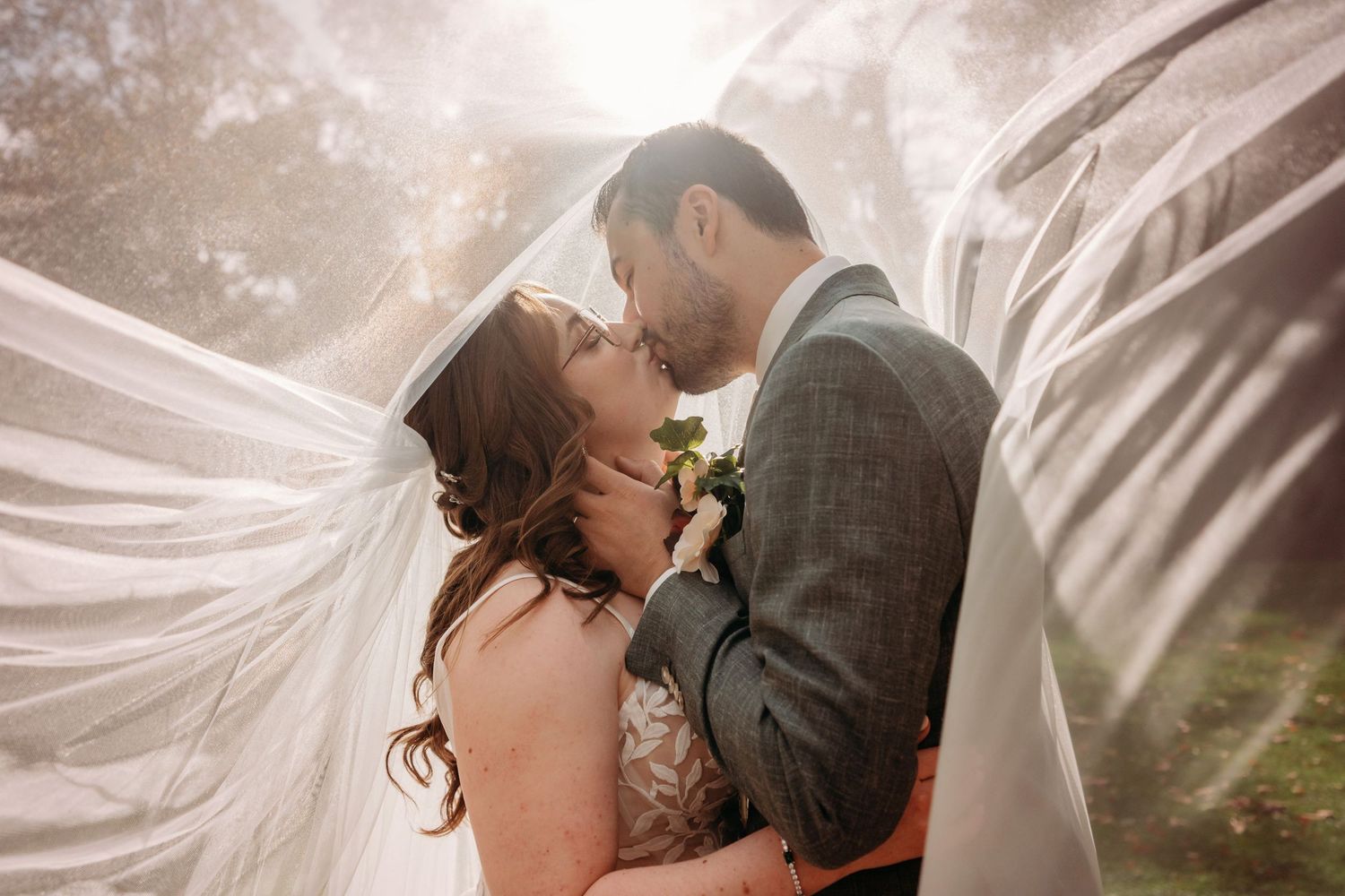 Bride and groom share a kiss beneath a flowing sheer veil in soft golden light, with the bride holding a small bouquet.