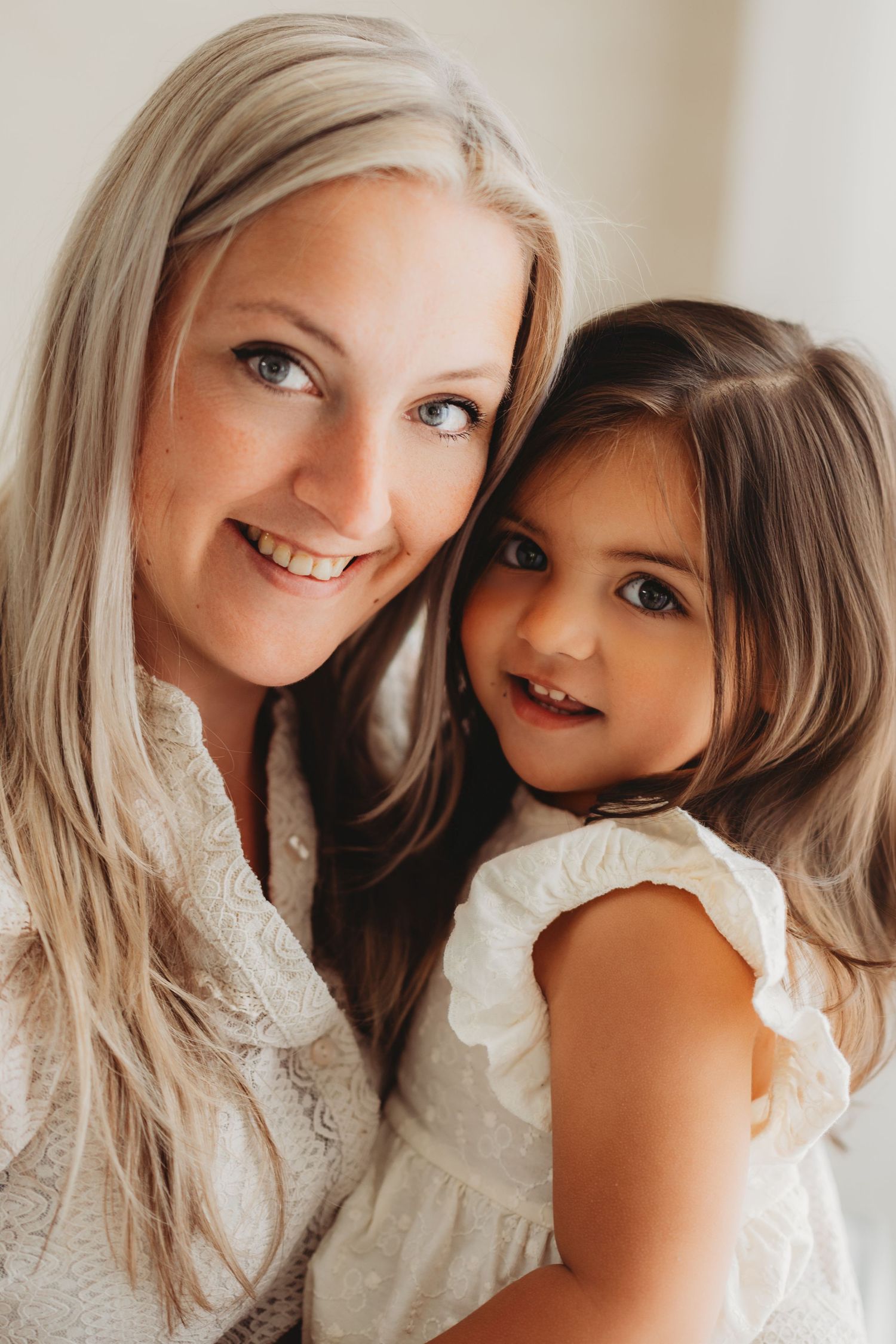 A smiling blonde woman with blue eyes holds a young brunette girl in a white dress, both posing for a portrait photo.