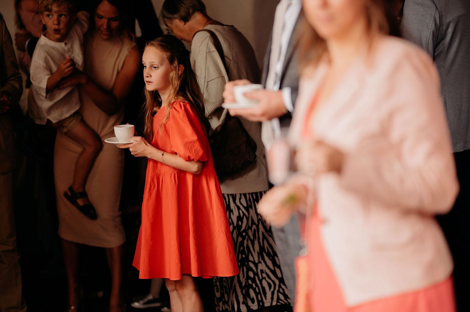 Young girl in red dress holding cup at wedding reception among guests in elegant venue.