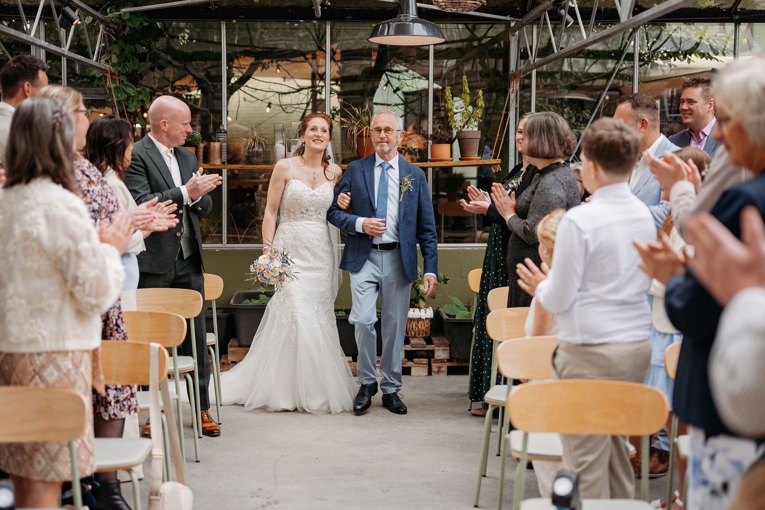 Bride walks down the aisle with her father as wedding guests applaud in a greenhouse venue.