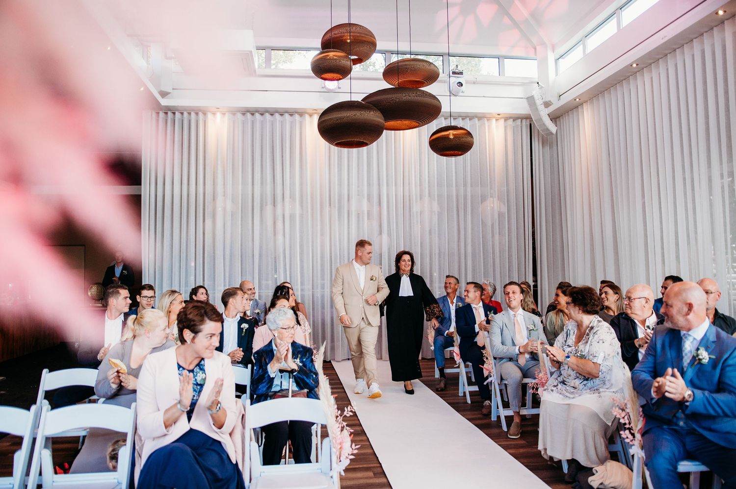 Trouwceremonie in moderne zaal met hanglampen en witte stoelen, bruidsfotografie IJM IJzereman Vught, Brabant.
