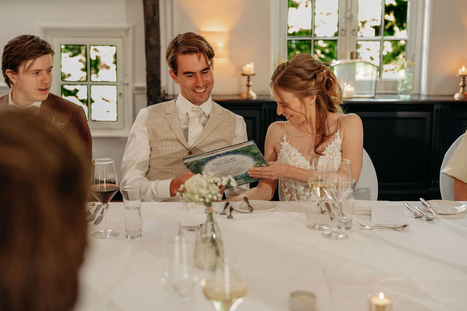 Bride and groom smiling together at wedding reception table, reviewing menu cards in elegant white-decorated venue.