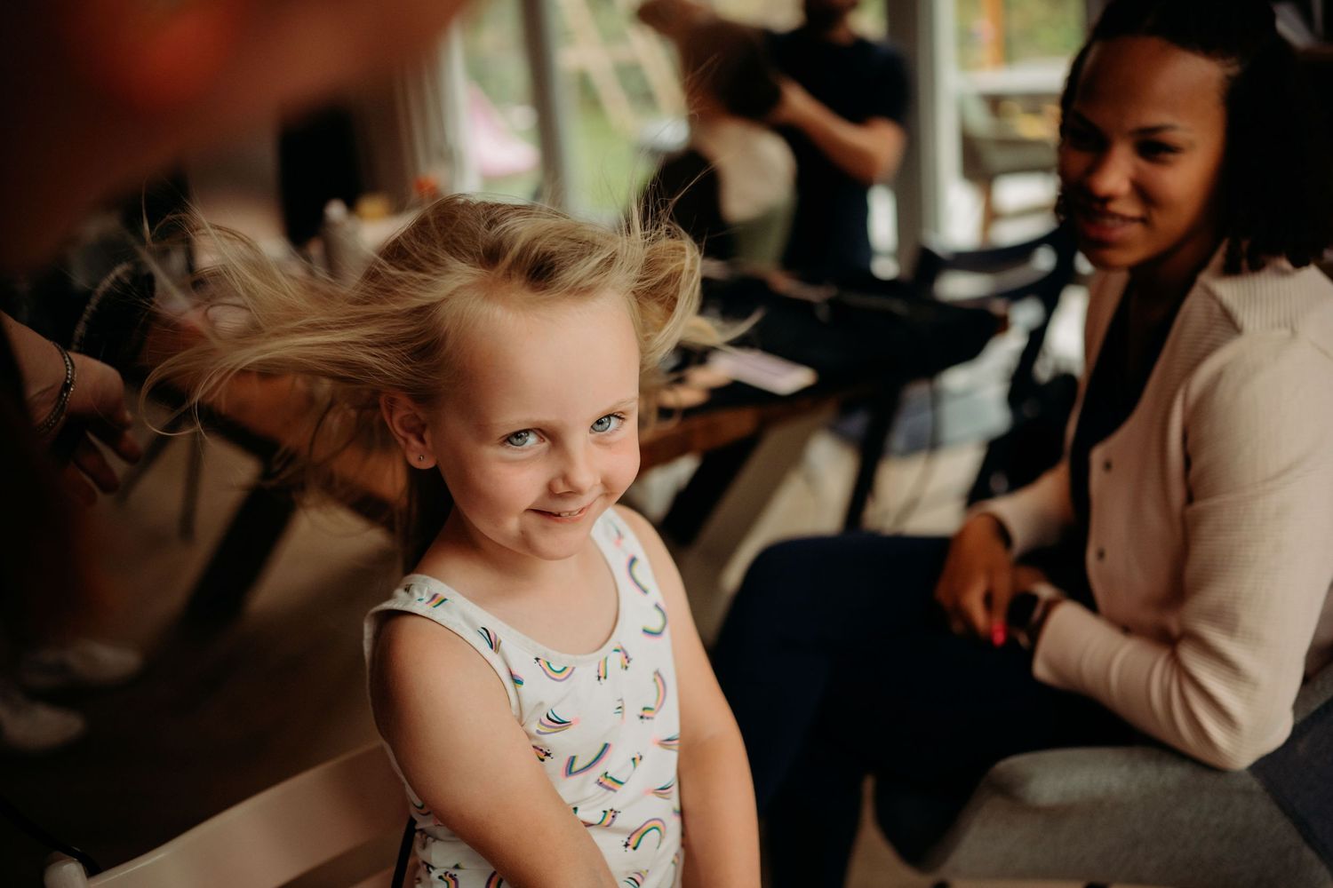 A young blonde girl smiles playfully with windswept hair while a woman sits smiling in the background.
