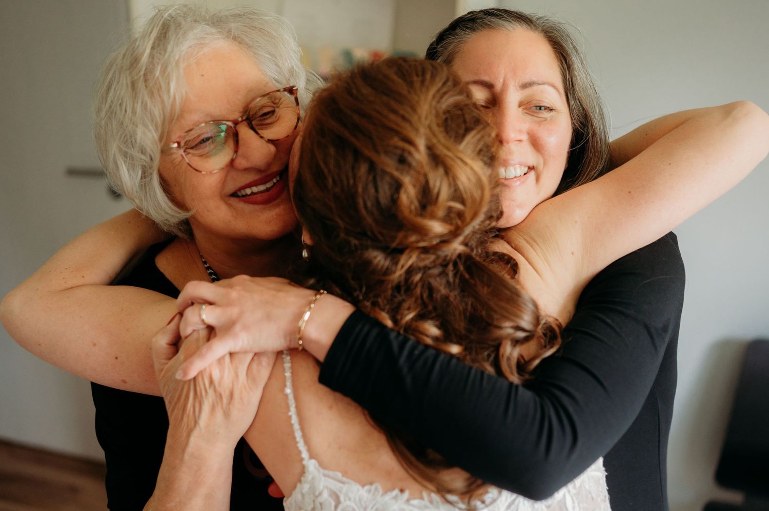 Two women embrace a bride in a warm group hug during wedding preparations.