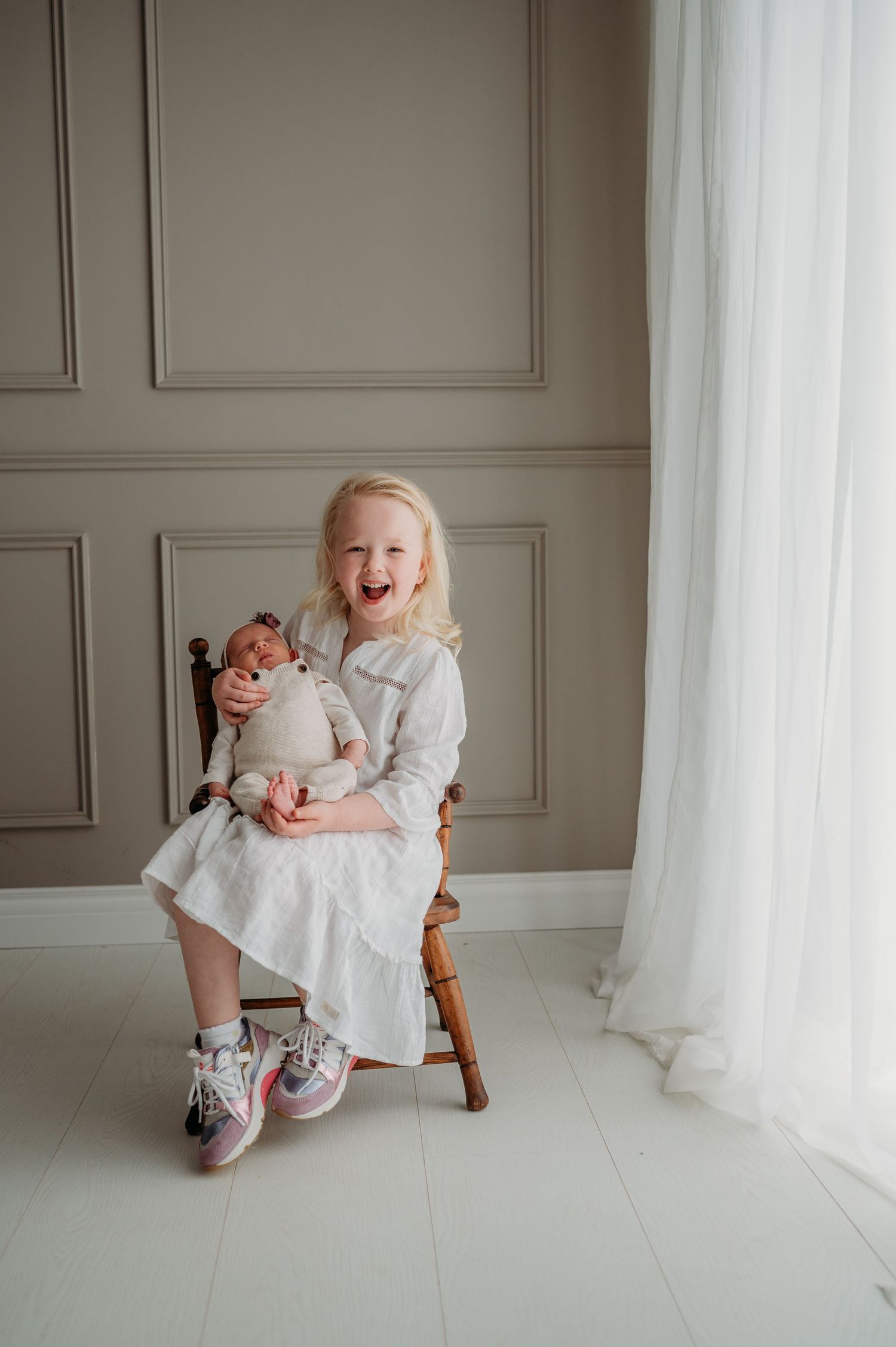 Lief portretsessie met witte jurk en schommelstoel tegen grijs paneel en witte gordijnen, familieshoot Brabant