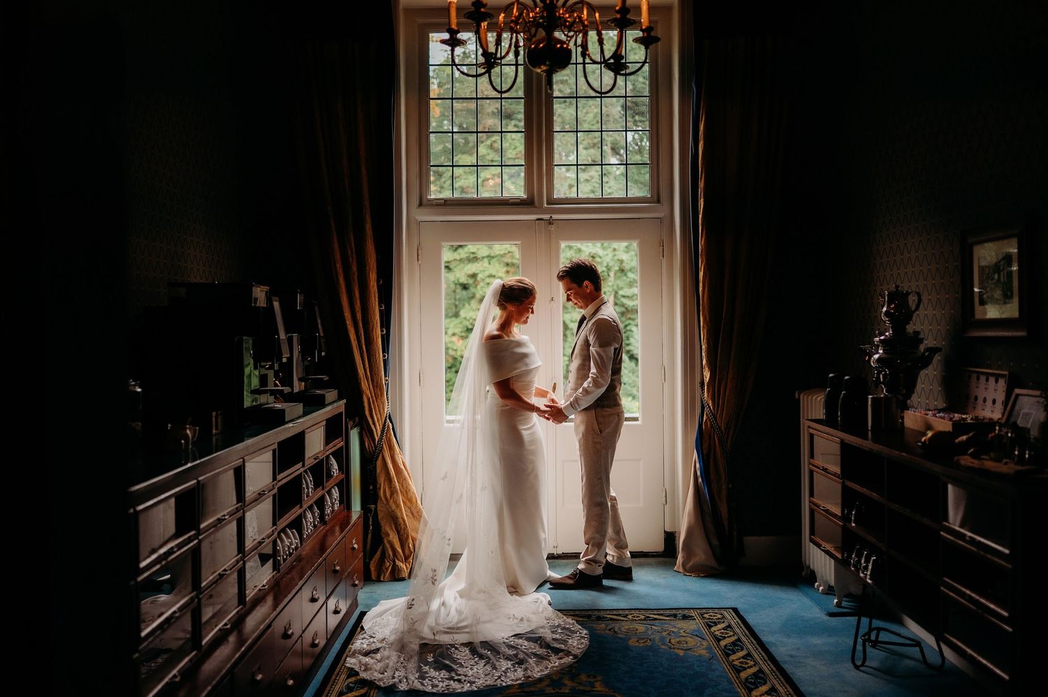 Bride and groom hold hands facing each other in a dark elegant room before a large window with golden drapes.