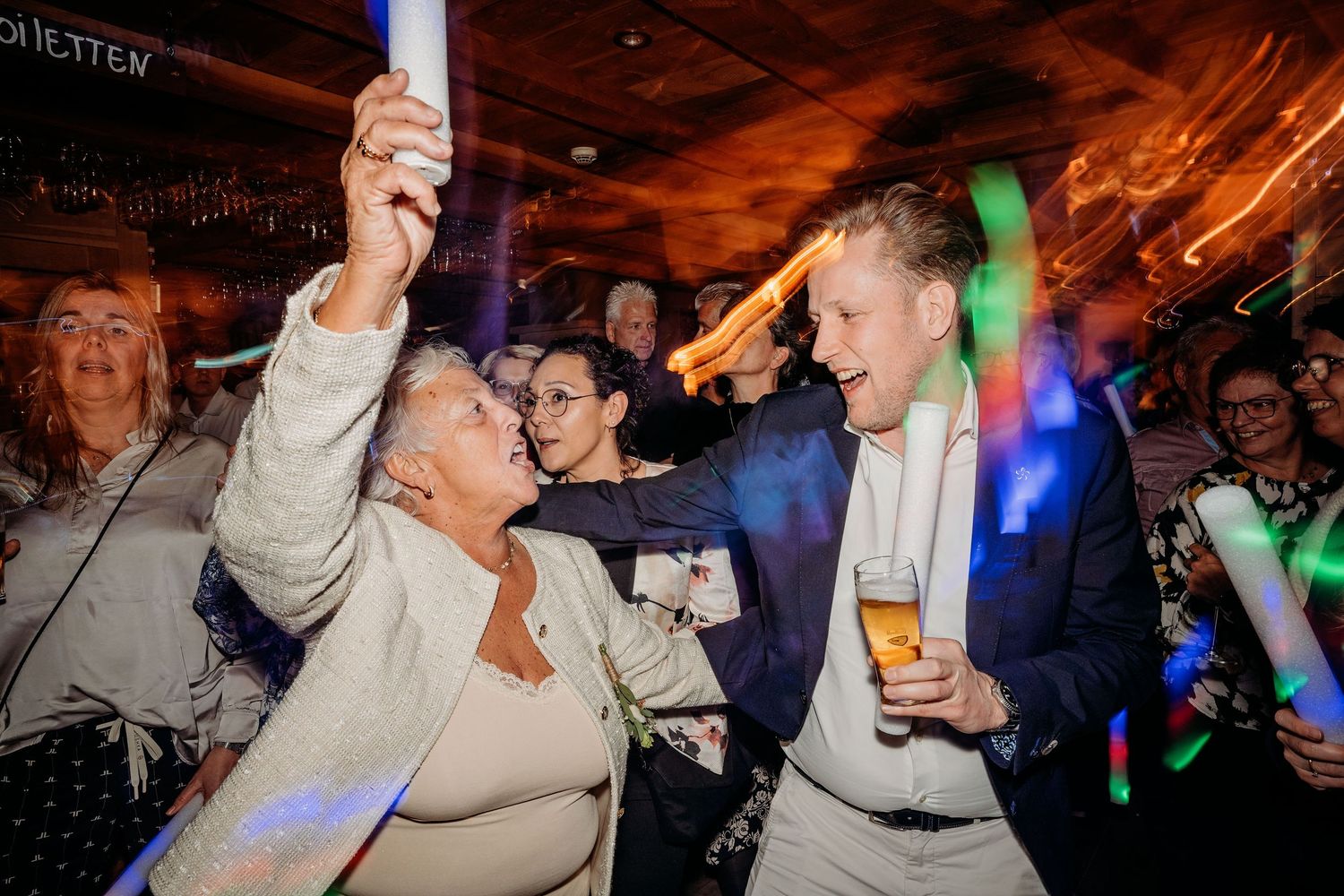 Gasten dansen uitbundig tijdens bruiloft feest in Brabant met kleurrijke verlichting, spontane fotografie