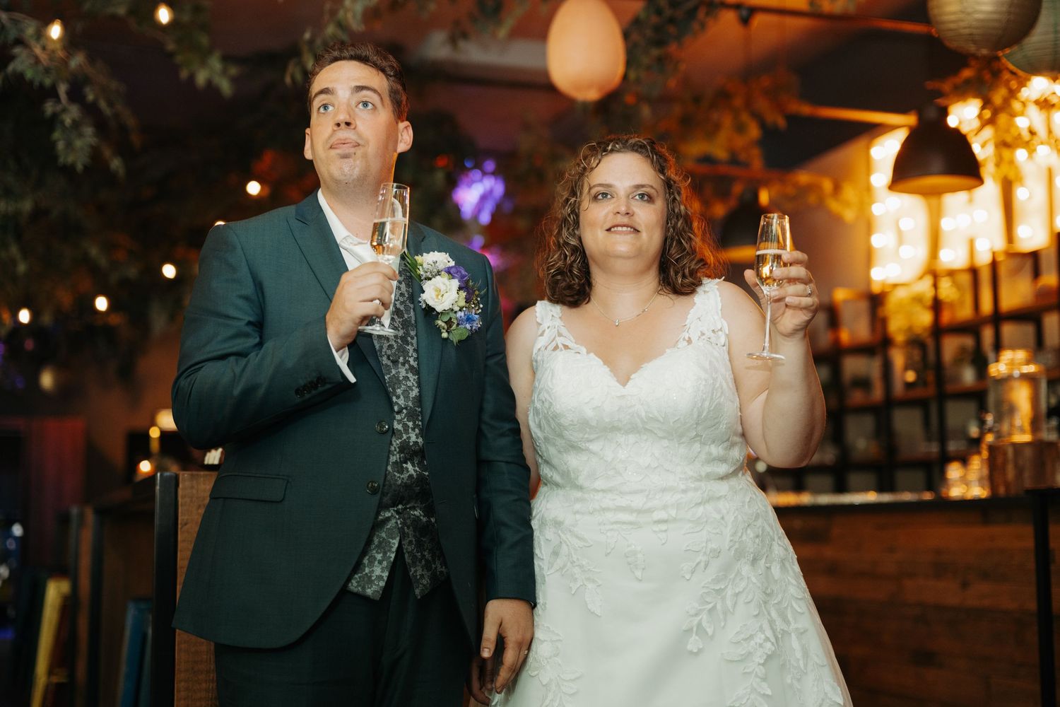 Bride in white lace gown and groom in teal suit raising champagne glasses during wedding reception toast.