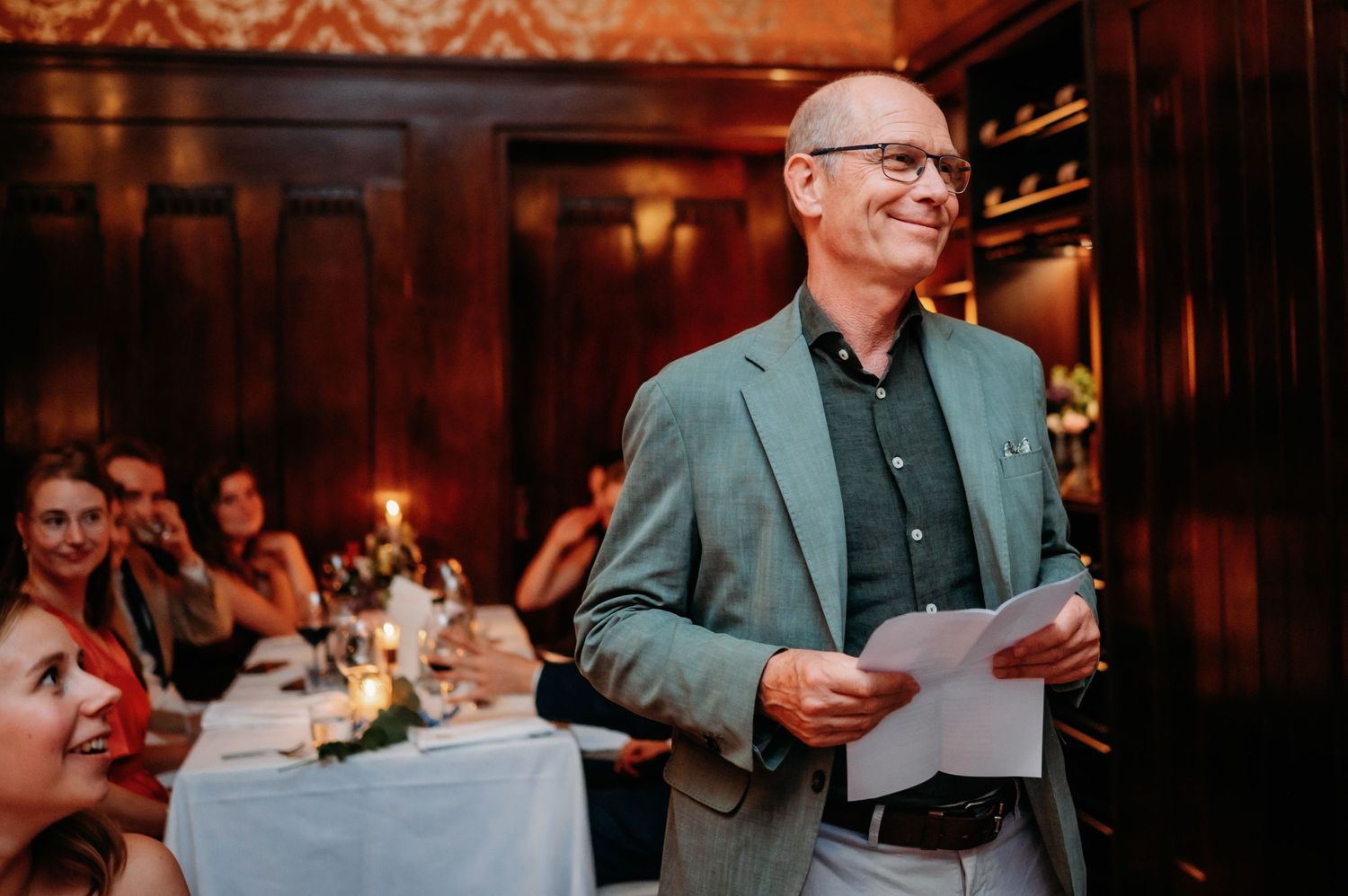 Man in green blazer giving a speech at an elegant candlelit wedding reception dinner.