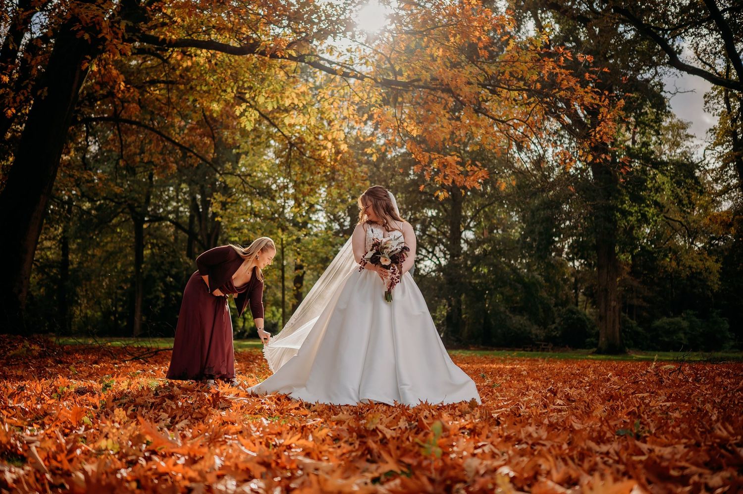 Bride in a flowing white wedding gown holding a bouquet while a bridesmaid adjusts her dress on a carpet of autumn leaves in a sunlit park.